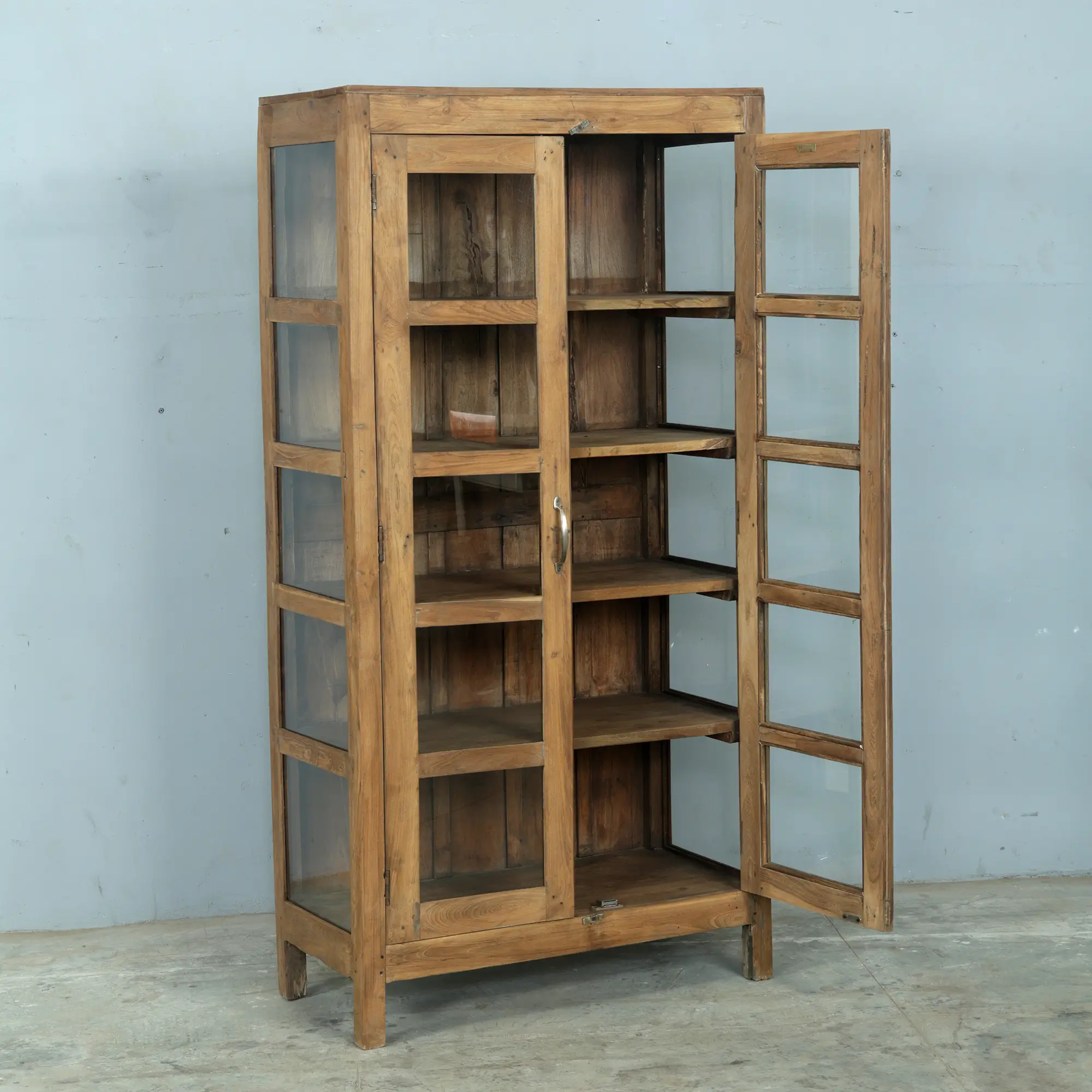 This teak and glass cabinet with brass handles was expertly crafted by hand from solid teak. It has five shelves and comes with glass doors and sides to let the light in. A beautiful piece of furniture to last forever.